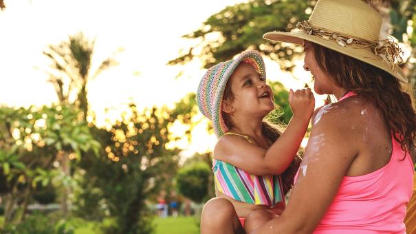 
		Happy mother sitting in the sunshine under a tree with her toddler and applying sunscreen
	